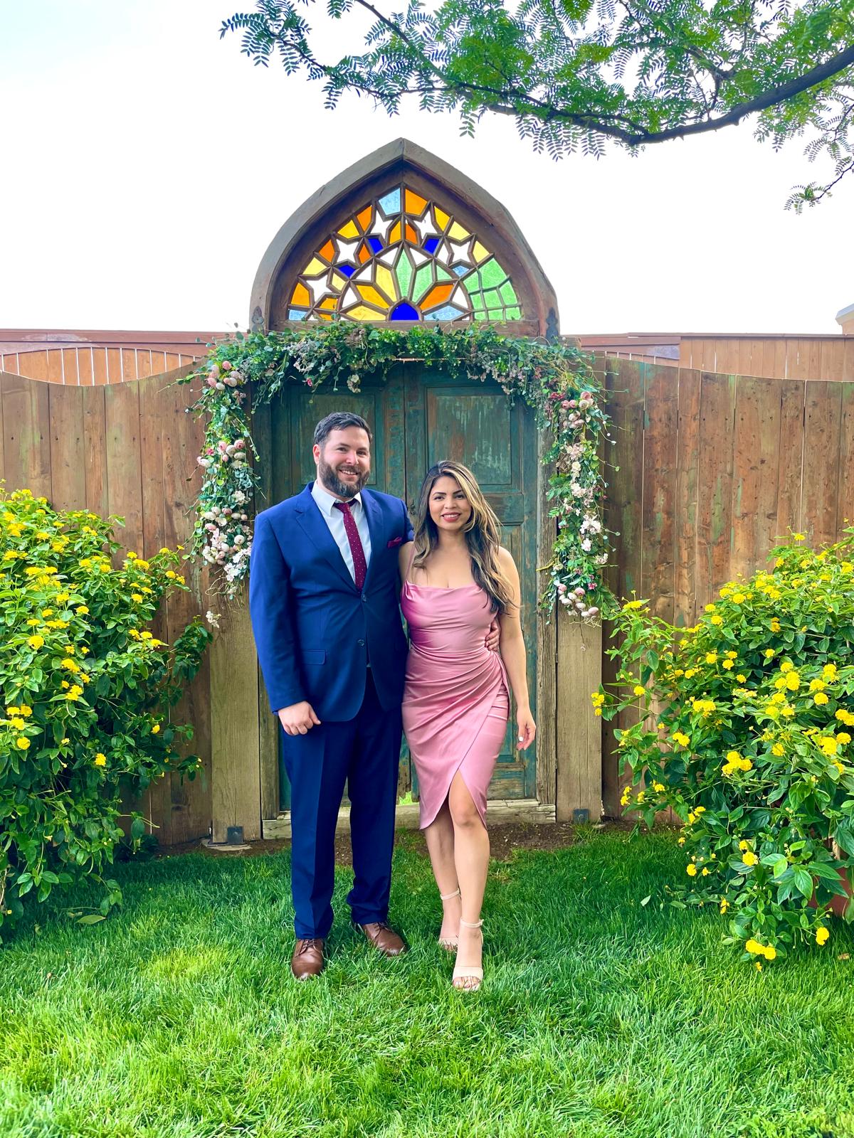 Travis in a blue suit and Karen in a pink dress beneath a flower-covered garden arch